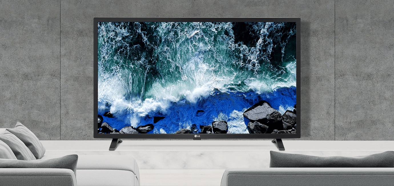 거실 공간에 배치된 LG TV가 테이블 위에 놓여 있으며, 화면에는 파도가 바위에 부딪히는 바다 풍경이 선명하게 표시됨. TV 앞에는 회색 톤의 소파와 쿠션이 배치되어 있고, 벽은 콘크리트 스타일의 모던한 디자인으로 구성됨.