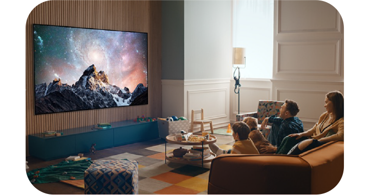 거실에서 가족이 소파에 앉아 벽걸이형 LG TV를 시청하는 모습. TV 화면에는 은하수가 배경으로 펼쳐진 산 풍경이 선명하게 표시됨. 거실에는 따뜻한 분위기의 조명과 장난감, 테이블 등이 배치되어 있어 아늑한 분위기를 연출함.