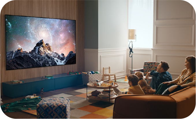 거실에서 가족이 소파에 앉아 벽걸이형 LG TV를 시청하는 모습. TV 화면에는 은하수가 배경으로 펼쳐진 산 풍경이 선명하게 표시됨. 거실에는 따뜻한 분위기의 조명과 장난감, 테이블 등이 배치되어 있어 아늑한 분위기를 연출함.