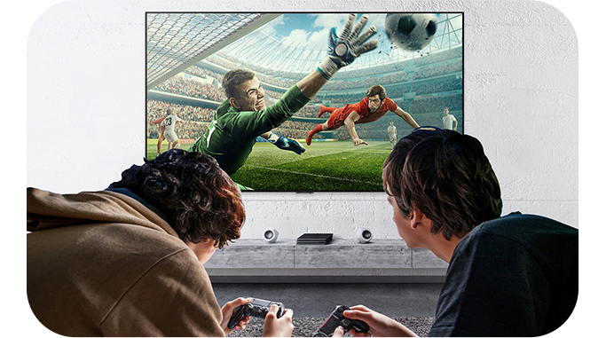 두 사람이 소파에 앉아 게임 컨트롤러를 들고 TV 화면을 보고 있다. TV에는 축구 경기 장면이 생생하게 재생되고 있으며, 골키퍼가 공을 막으려는 순간을 담고 있다. 게임의 몰입감을 강조하는 모습이다.