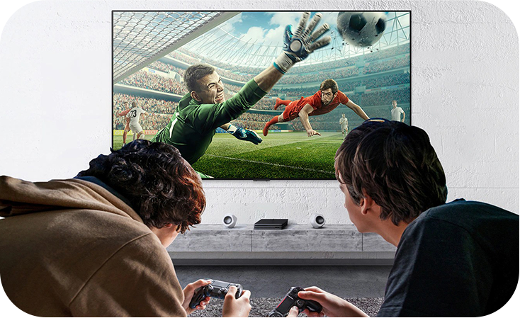 두 사람이 소파에 앉아 게임 컨트롤러를 들고 TV 화면을 보고 있다. TV에는 축구 경기 장면이 생생하게 재생되고 있으며, 골키퍼가 공을 막으려는 순간을 담고 있다. 게임의 몰입감을 강조하는 모습이다.