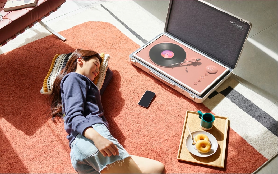 햇살이 드는 거실 바닥에 러그를 깔고 한 여성이 편안하게 누워 있다. 그녀 옆에는 LG StanbyME Go가 열려 있으며, 화면에는 빈티지한 레코드 플레이어 인터페이스가 표시되어 있다. 스마트폰이 바닥에 놓여 있고, 옆에는 도넛과 커피가 담긴 나무 쟁반이 있다. 따뜻한 분위기 속에서 음악을 감상하며 여유로운 시간을 보내는 모습이다.