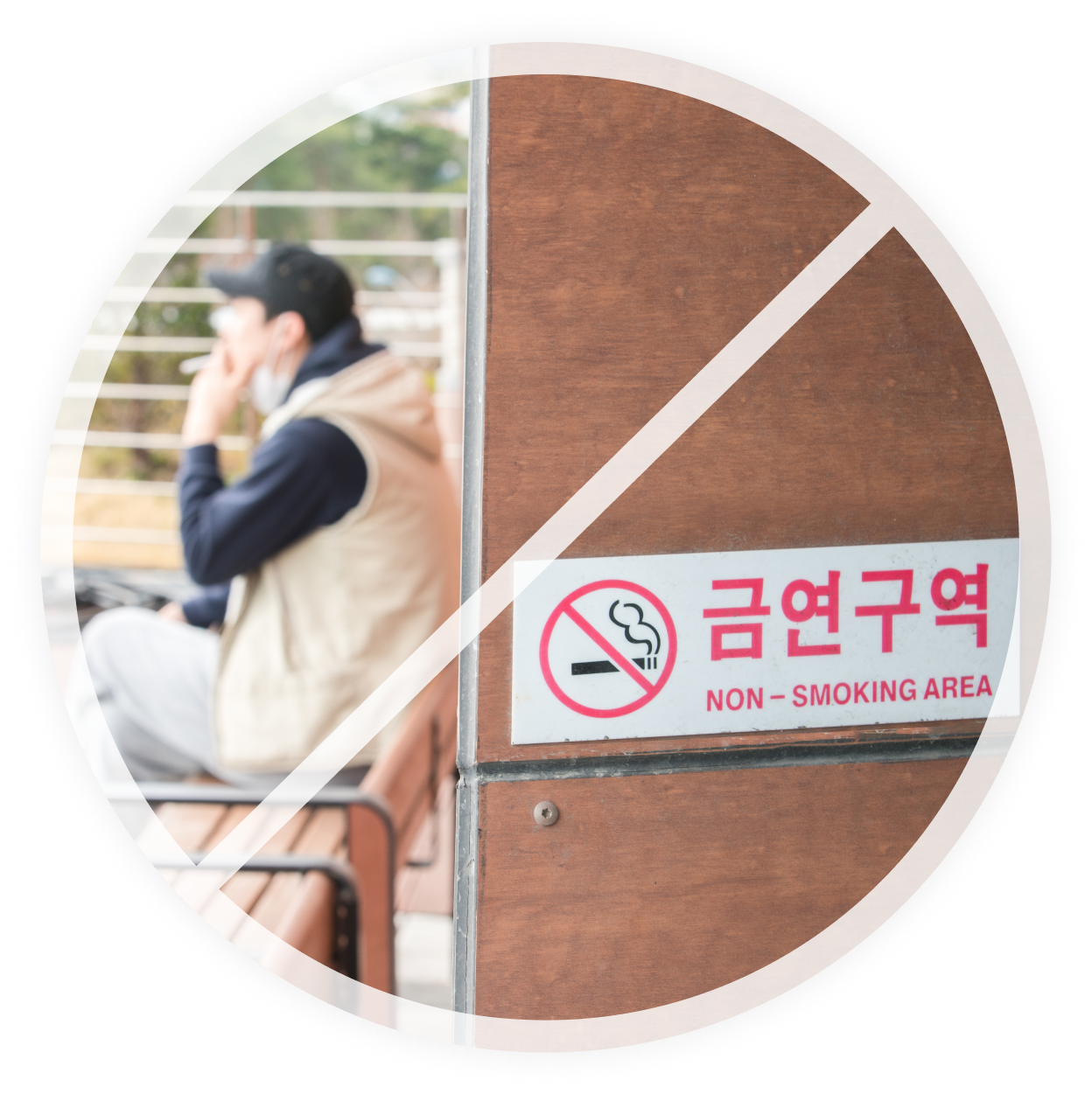  '금연구역'이라는 표시와 금연 마크가 나무 벽에 부착된 모습. 배경에는 한 남성이 벤치에 앉아 담배를 피우는 모습이 흐릿하게 보임.