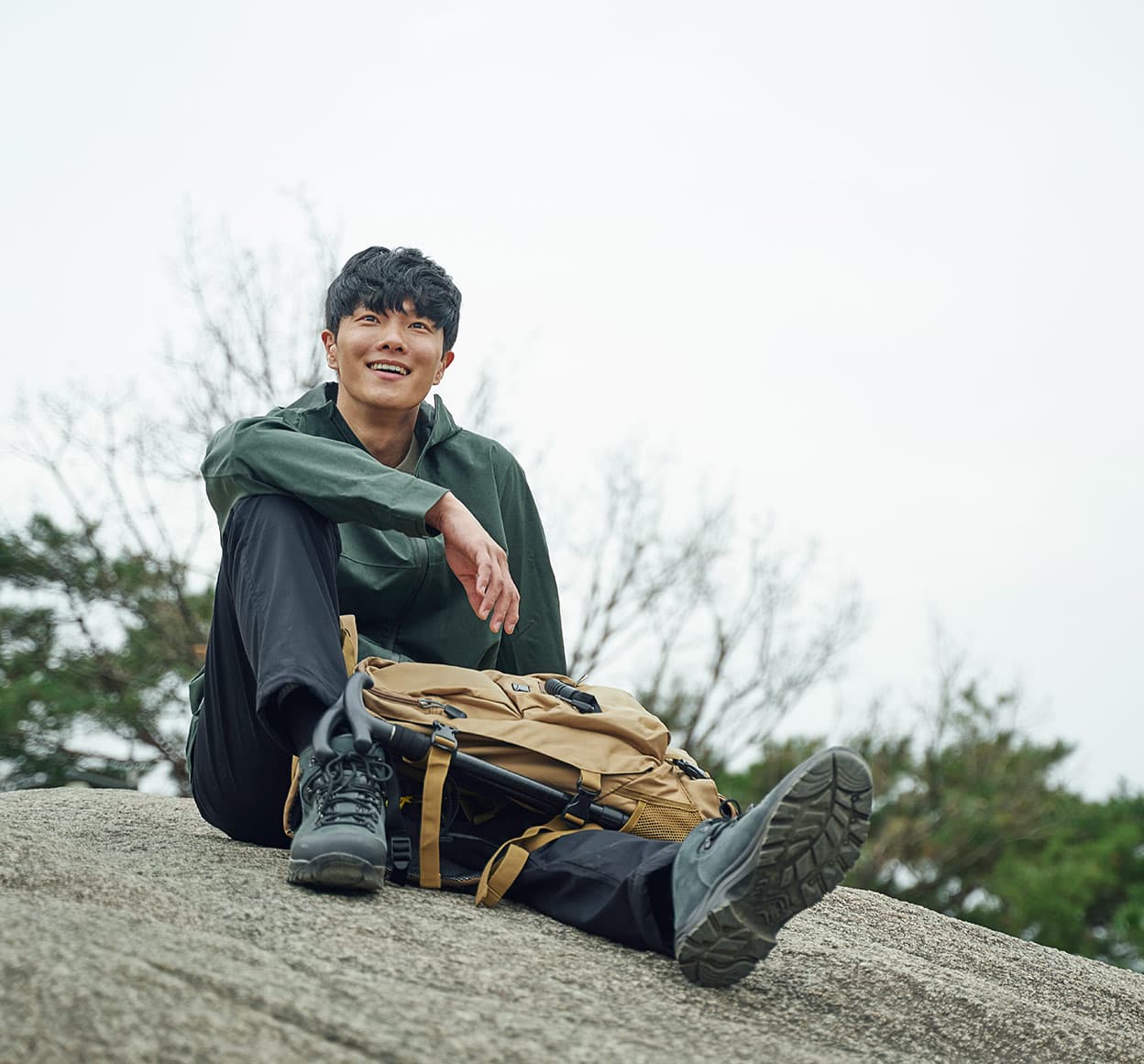 짙은 녹색 재킷과 검은색 등산화를 착용한 남성이 바위 위에 앉아 환하게 웃고 있음. 옆에는 베이지색 등산 배낭과 접이식 스틱이 놓여 있음.