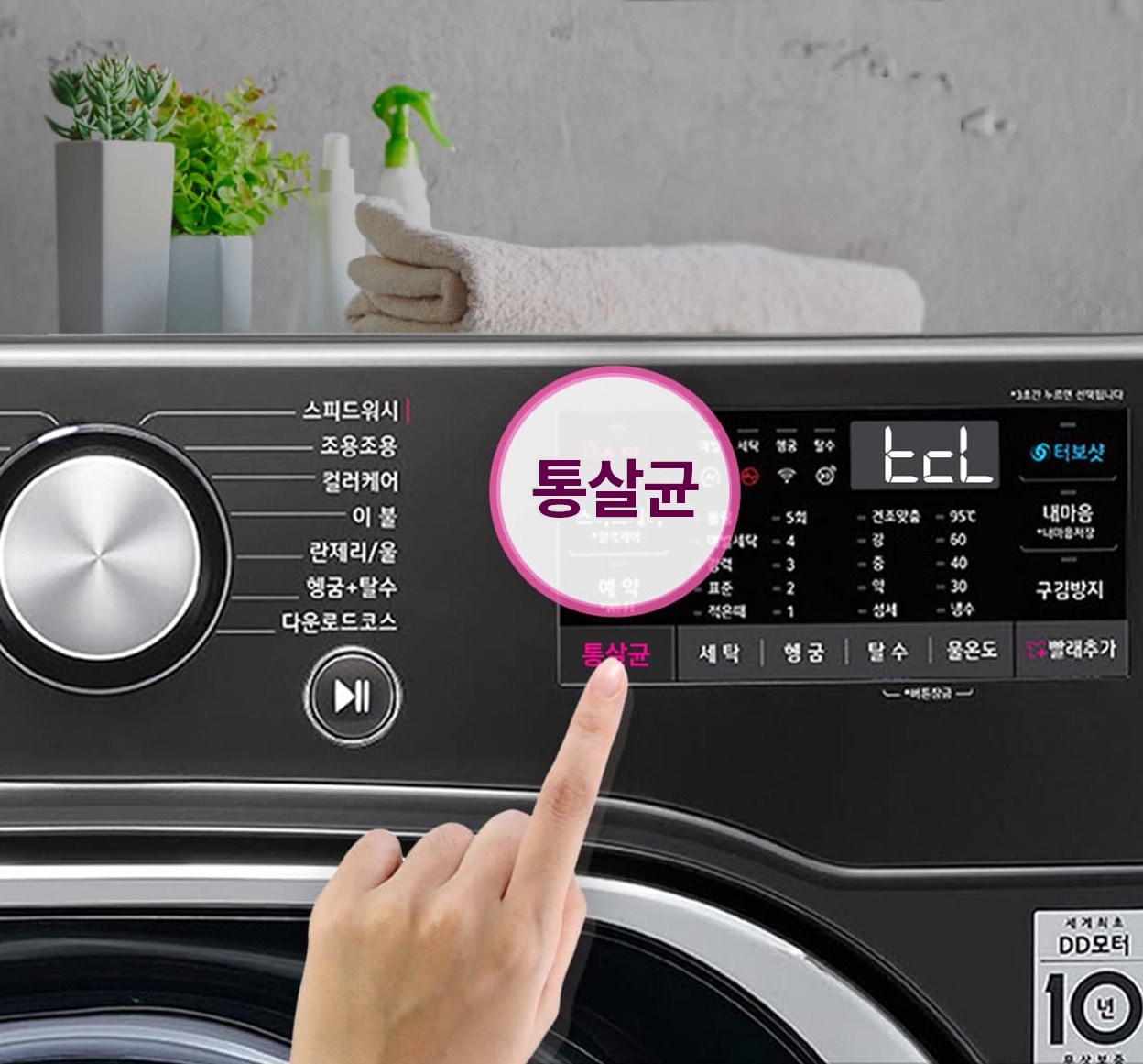 검은색 세탁기 조작 패널을 손가락으로 터치하고 있음. 패널에는 다양한 세탁 옵션이 표시돼 이고, 통살균 기능이 강조되어 있음. 세탁기 위에는 접힌 수건과 화분, 세제가 있음. 제품 기능 및 기능 명은 모델별 상이 문구 표시됨.