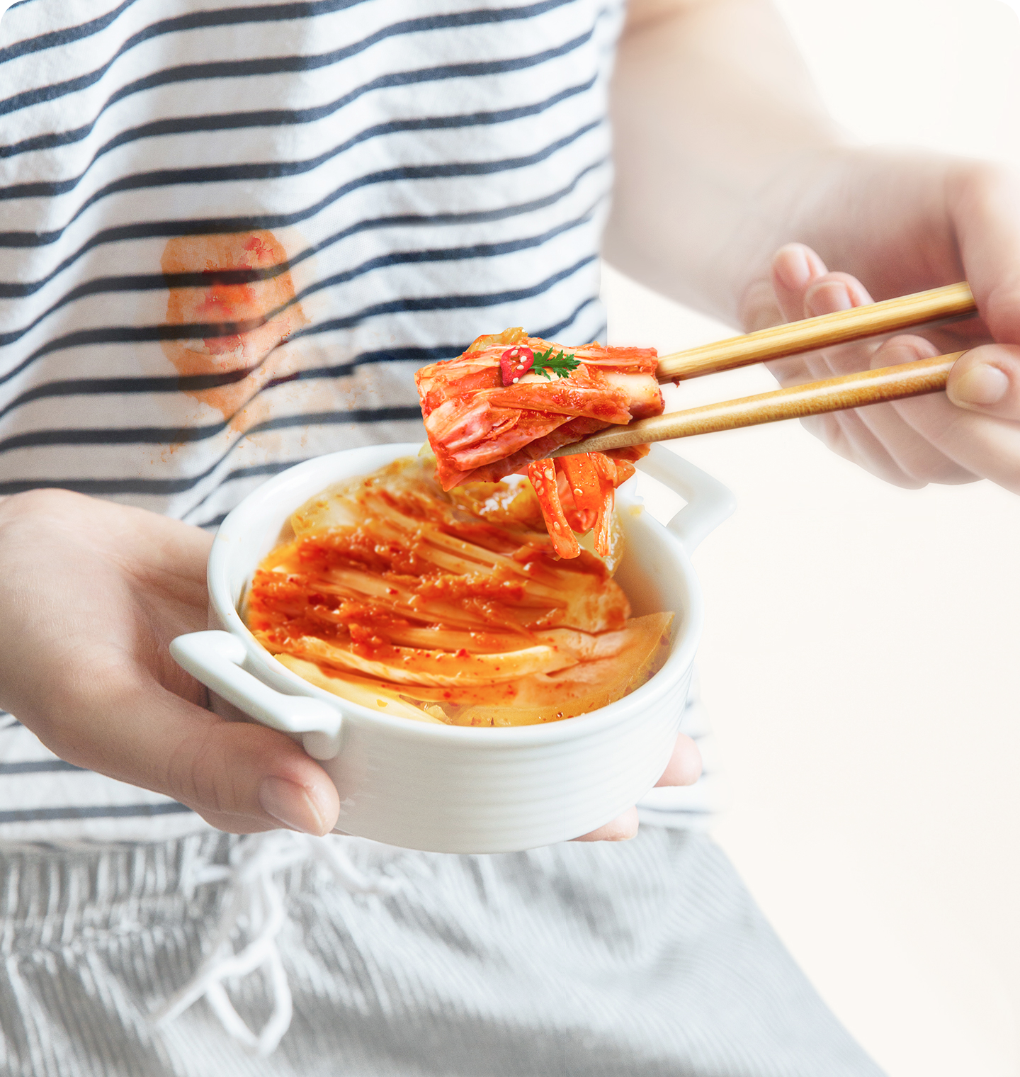 줄무늬 티셔츠를 입은 사람이 한 손으로 흰색 김치 그릇을 들고, 다른 손으로 젓가락을 사용해 김치를 집어 먹고 있음. 옷에는 김치 국물 얼룩이 있음.