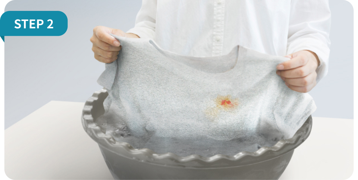 step2. 김치 얼룩이 묻은 밝은 회색 티셔츠를 들고 있는 손과 물에 담그는 모습이 보임.
