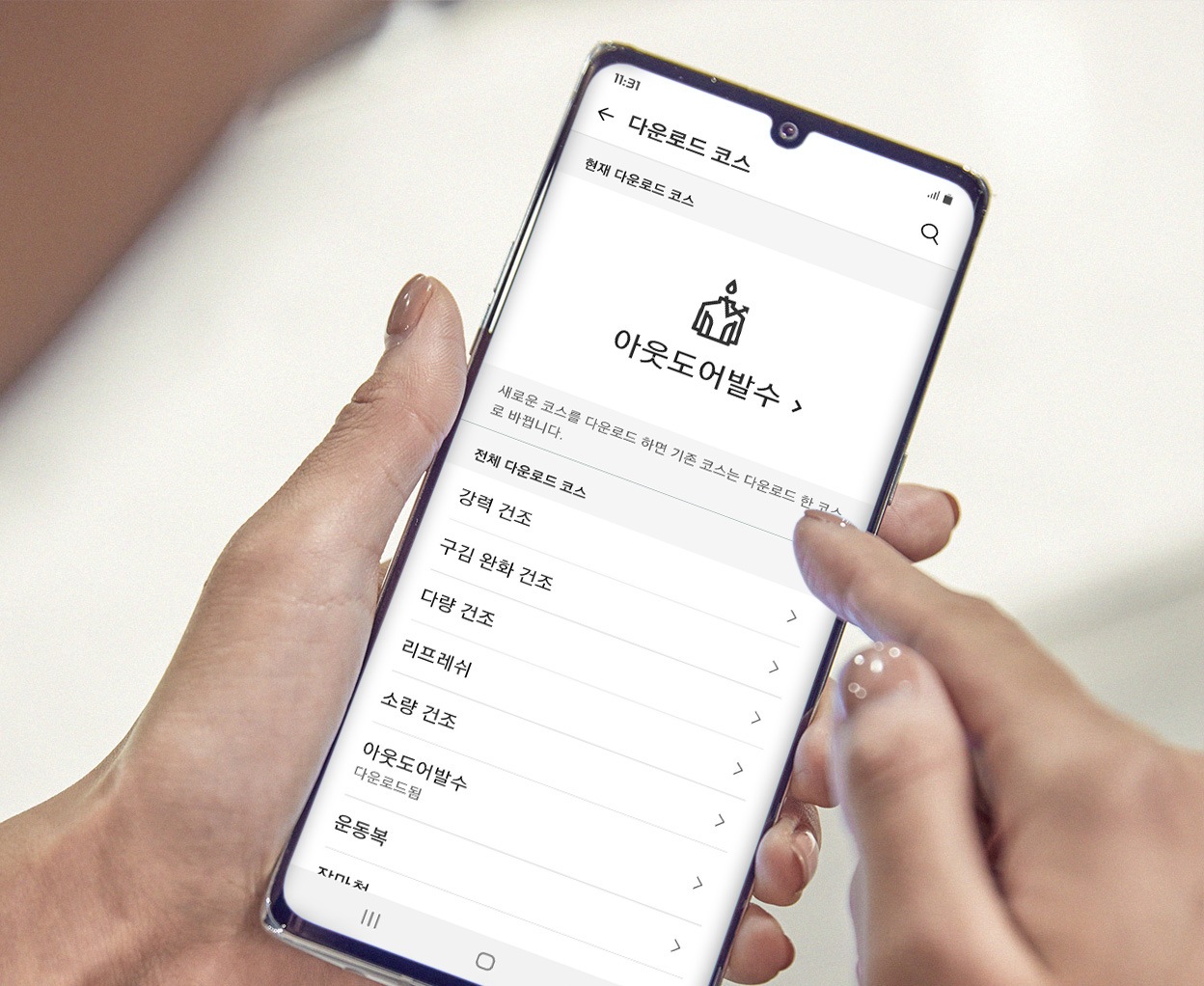스마트폰 화면에서 건조기 다운로드 코스를 설정하는 모습, 사용자가 아웃도어헹굼 옵션을 선택 중임. 손끝이 화면을 터치하며 기능을 조정하는 장면.