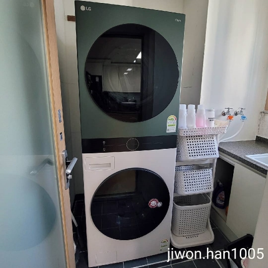 베란다 공간에 세탁기와 건조기가 세로로 배치되어 있고, 옆에는 세탁 용품을 보관하는 선반이 정리되어 있으며, 하얀색 커튼이 걸려 있음