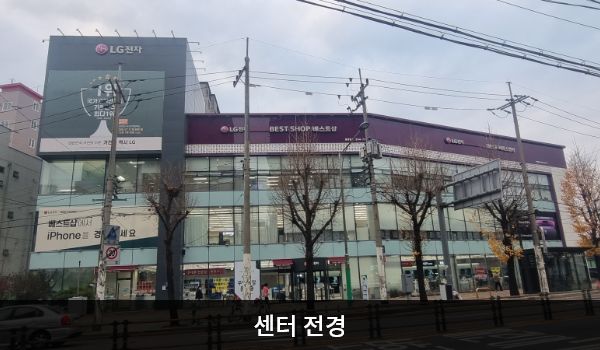 은평서비스센터의 서비스센터 건물은 왼쪽 부분만 타워형으로 높이가 더 높은 특징이 있습니다. 벽면에 큰 창문이 많습니다. 건물 위 간판에 흰색 글씨, 분홍색 바탕으로 'LG전자 BEST SHOP 베스트샵', 'BEST 서비스센터'라고 쓰여 있습니다.