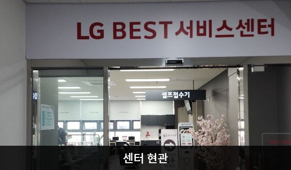 이천서비스센터의 센터 현관 위쪽에 'LG BEST 서비스센터'라는 문구가 적혀 있으며, 그 아래에 유리로 된 자동문이 있습니다. 문을 열고 들어가면 앞에 셀프 접수기가 있습니다.