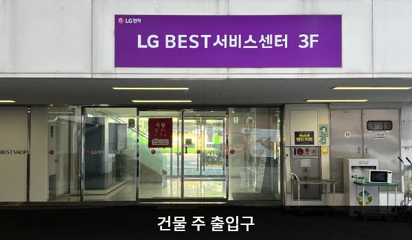 남인천서비스센터의 건물 주 출입구 위에는 보라색 바탕에 'LG BEST 서비스센터 3F'라는 문구가 크게 적혀 있습니다. 자동문으로 되어 있으며 오른쪽 문 안쪽에 문 열림 버튼이 있습니다. 문 양옆으로는 유리창이 있습니다.