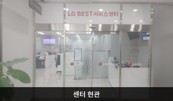 군산서비스센터의 센터 현관 위쪽에 'LG BEST 서비스센터'라는 문구가 적혀 있으며, 그 아래에 자동문이 있습니다. 문을 열고 들어가면 앞에 셀프 접수기가 있습니다.