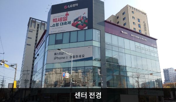 관악서비스센터의 서비스센터 건물은 다각형이며, 전체가 유리창으로 되어 있습니다. 건물 꼭대기 간판에 흰색 글씨, 분홍색 바탕으로 'LG전자 BEST SHOP & 서비스센터'라고 쓰여 있습니다.