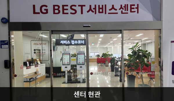 일산서비스센터의 센터 현관 위쪽에 'LG BEST 서비스센터'라는 문구가 적혀 있으며, 그 아래에 유리로 된 자동문이 있습니다. 문을 열고 들어가면 앞에 셀프 접수기가 있습니다.