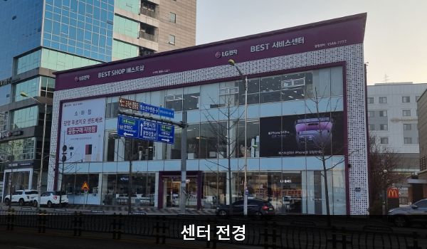 광명서비스센터의 서비스센터 건물은 가로로 길고 유리창이 많습니다. 건물 위 간판에 흰색 글씨, 분홍색 바탕으로 'LG전자 BEST SHOP 베스트샵', 'LG전자 BEST 서비스센터'라고 쓰여 있습니다.