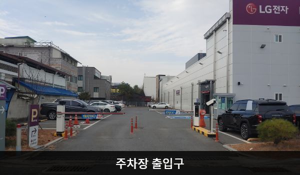 충주서비스센터의 건물 뒤편, 주 출입구 앞쪽에 지상 주차장이 있습니다. 'P'라는 글자가 크게 적힌 보라색 입간판 우측으로 주차장 출입구가 있습니다.