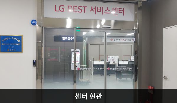 충주서비스센터의 센터 현관 위쪽에 'LG BEST 서비스센터'라는 문구가 적혀 있으며, 그 아래에 유리로 된 자동문이 있습니다. 문을 열고 들어가면 앞에 셀프 접수기가 있습니다.