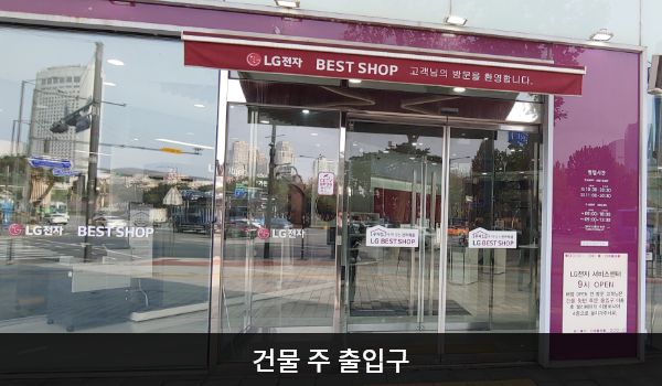 송파서비스센터의 건물 주 출입구 위에는 붉은색 차양막이 있으며, 그 아래에 자동문이 있습니다. 자동문을 열고 들어가면 여닫을 수 있는 유리문이 하나 더 있습니다.