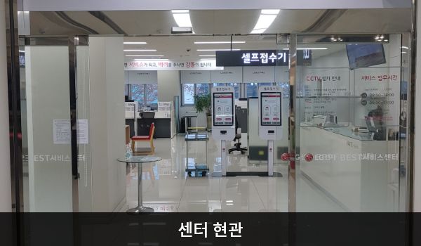 안산서비스센터의 센터 현관 위쪽에 'LG BEST 서비스센터'라는 문구가 적혀 있으며, 그 아래에 유리로 된 자동문이 있습니다. 문을 열고 들어가면 정면에 셀프 접수기가 있습니다.