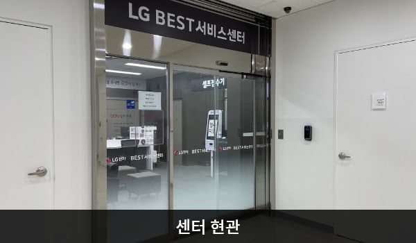 아산서비스센터의 센터 현관 위쪽에 'LG BEST 서비스센터'라는 문구가 적혀 있으며, 그 아래에 유리로 된 자동문이 있습니다. 문을 열고 들어가면 정면 우측에 셀프 접수기가 있습니다.