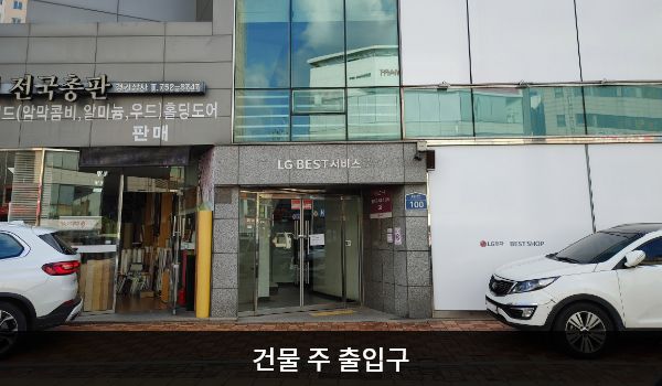 성남서비스센터의 건물 출입구는 2개로, 도로 쪽과 주차장 쪽에 있습니다. 도로 쪽 출입구는 회색 돌벽으로 둘러싸여 있으며, 출입문 위에 'LG BEST 서비스'라고 쓰여 있습니다. 양쪽 유리문 안쪽에 긴 손잡이가 있습니다.