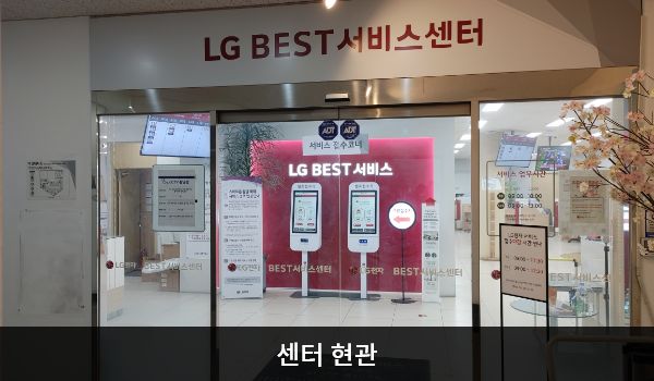 성남서비스센터의 센터 현관 위쪽에 'LG BEST 서비스센터'라는 문구가 적혀 있으며, 그 아래에 유리로 된 자동문이 있습니다. 문을 열고 들어가면 정면에 셀프 접수기가 있습니다.