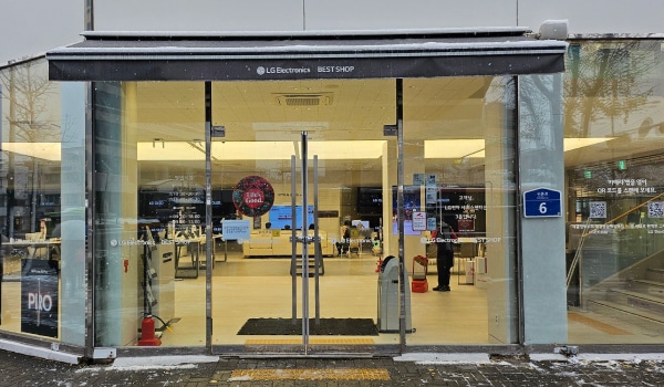 홍대역서비스센터의 건물 주 출입구 위 검은색 차양막에 작은 글씨로 'LG Electronics BEST SHOP'이라고 적혀 있습니다. 유리문으로 되어 있으며, 양쪽 문 안쪽에 긴 손잡이가 있습니다.