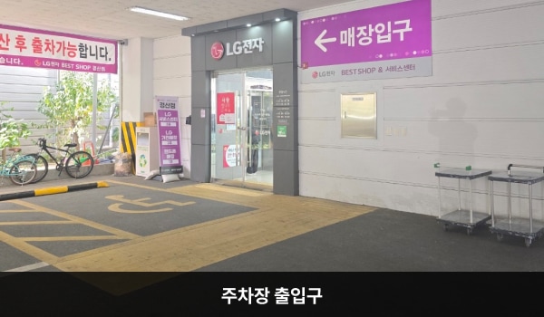 경산서비스센터의 건물 왼쪽 끝에 주차장 출입구가 있습니다. 입구 위 간판에 'LG전자 고객전용주차장'이라고 쓰여 있습니다. '매장 입구'라고 쓰인 안내문 옆 여닫이문이 건물 출입구이며 바로 왼쪽에 엘리베이터가 있습니다.