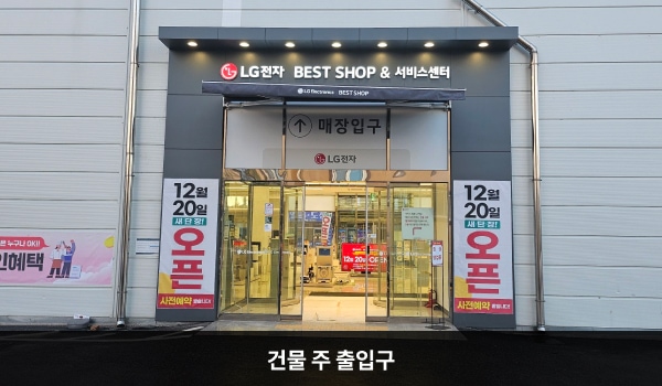 논산서비스센터의 건물 주 출입구 붉은색 차양막 위에 '휴대폰 전문점'이라고 적힌 돌출된 간판이 있습니다. 자동문 가운데 문 열림 버튼이 있습니다.