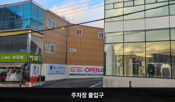 논산서비스센터의 건물 왼쪽에 주차장 출입구가 있습니다. 출입구 왼쪽에 'P'라고 적힌 보라색 입간판이 있으며, 그 옆쪽 벽에는 '매장입구'라고 적힌 보라색 간판이 붙어 있습니다.