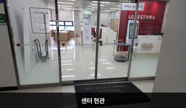 논산서비스센터의 센터 현관 위쪽에 'LG BEST 서비스센터'라는 문구가 적혀 있으며, 그 아래에 유리로 된 자동문이 있습니다. 문을 열고 들어가면 앞에 셀프 접수기가 있습니다.