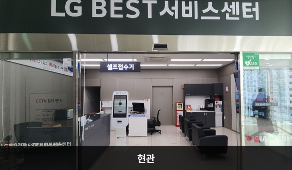 김천서비스센터의 엘리베이터 정면에 센터 현관이 있습니다. 유리로 된 자동문 위쪽 큰 검은색 간판에 'LG BEST 서비스센터'라는 문구가 적혀 있습니다. 들어오시면 앞에 셀프 접수기가 있고 왼쪽에는 부품 판매 창구, 오른쪽에는 고객 대기실이 있습니다.