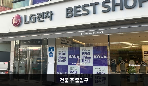 홍성서비스센터의 건물 주 출입구 위 흰색 간판에 'LG전자 BEST SHOP'이라는 문구가 적혀 있습니다. 양 유리문 안쪽에 긴 손잡이가 있습니다. 서비스센터는 3층입니다.