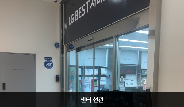 강동서비스센터의 센터 현관 위쪽에 'LG BEST 서비스센터'라는 문구가 적혀 있으며, 그 아래에 유리로 된 자동문이 있습니다. 문을 열고 안쪽으로 들어가면 정면에 셀프 접수기가 있습니다.
