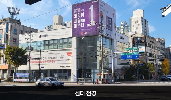 노원서비스센터의 서비스센터 건물은 한 부분만 타워형으로 높이가 더 높은 특징이 있습니다. 벽면에 큰 창문이 많습니다. 건물 위 간판에 분홍색 바탕, 흰색 글씨로 'LG전자 BEST SHOP & 서비스센터'라고 쓰여 있습니다. 대로 앞 베스트샵과 연결된 출입문 앞에 점자블록이 있습니다.