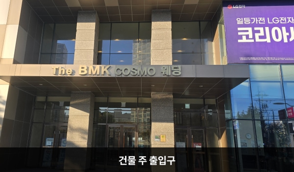 서대전서비스센터의 1층에는 LG BEST SHOP이 있고, 그 옆에 2층 서비스센터로 진입하실 수 있는 전체 건물 출입구가 있습니다. 출입구 위에는 'The BMK COSMO 웨딩'이라고 적힌 간판이 있습니다. 2개의 큰 기둥 사이에 위치한 여닫이문이 있습니다.