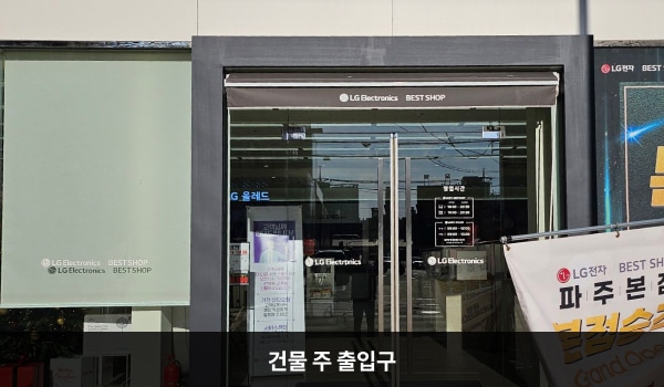 파주서비스센터의 건물 정면에 주 출입구가 있습니다. 출입구 위쪽에는 검은색 차양막이 있으며, 문 앞에 점자블록이 있습니다. 양 유리문 안쪽에는 긴 손잡이가 있습니다.