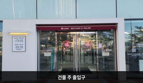 유성서비스센터의 건물 가운데에 여닫이문으로 된 건물 출입구가 있고, 문 앞에 점자블록이 있습니다. 양쪽 유리문에 긴 손잡이가 있습니다. 서비스센터는 건물 4층에 있습니다.