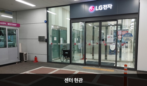 미사본점서비스센터의 센터 현관 위에는 검은색 바탕에 'LG전자'라는 문구가 적혀 있습니다. 자동문 앞에 점자블록이 있으며, 자동문을 통해 들어가면 유리문이 하나 더 있습니다.