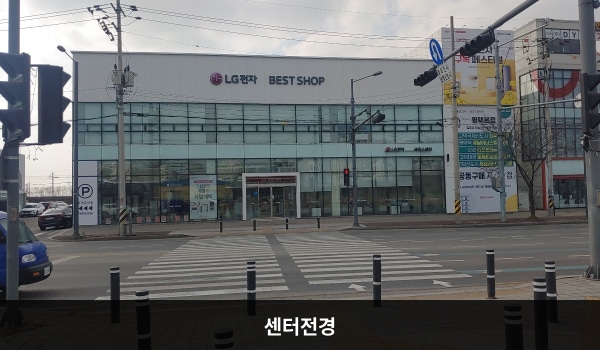 평택서비스센터의 서비스센터 건물은 가로로 길고 흰색이며 창문이 많습니다. 건물 2층 간판에 검정색 글씨로 'LG전자 BEST SHOP'이라고 쓰여 있습니다.