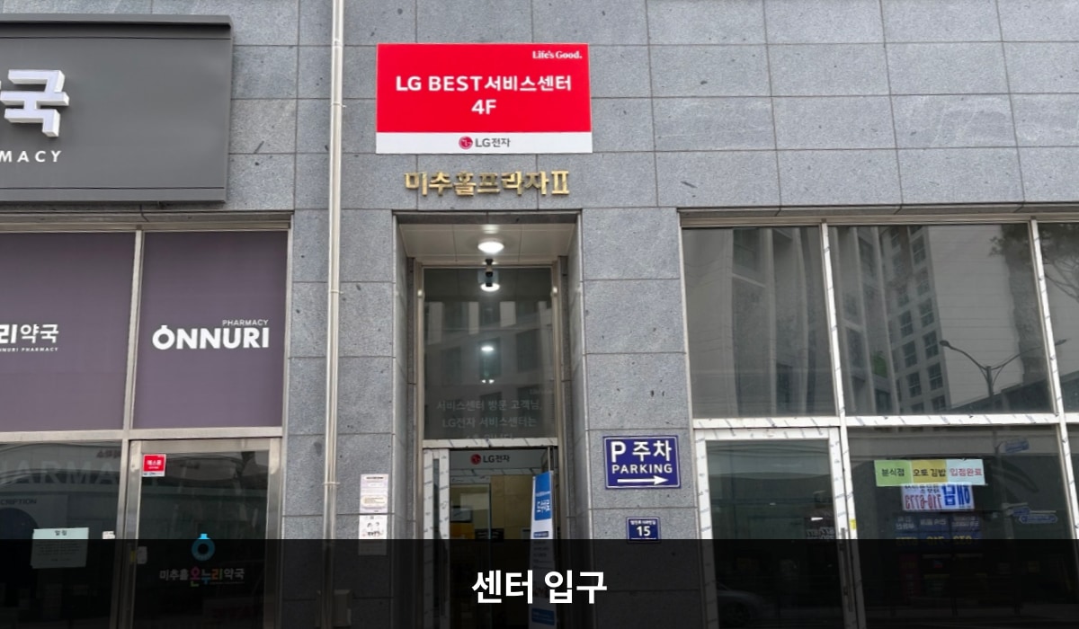 남인천서비스센터의 건물 정면 중앙에 주 출입구가 있습니다. 유리문으로 되어 있으며, 문 왼쪽에 긴 손잡이가 있습니다. 문 위쪽 벽면에는 'LG BEST 서비스센터 4F', '미추홀프라자Ⅱ'라는 문구가 적혀 있습니다.