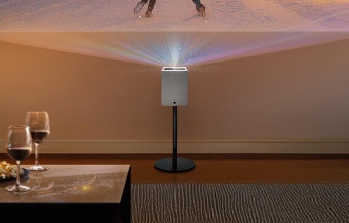 스탠드 위에 설치된 큐브 모양의 프로젝터가 정면 벽에 영상을 투사하고 있으며, 전경에는 와인잔과 테이블이 보입니다.