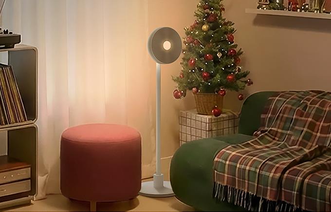 방 중앙에는 원형 조명 헤드를 가진 흰색 스탠드 조명이 서 있고, 그 옆에는 분홍색 스툴과 체크무늬 담요가 덮인 초록색 안락의자가 있습니다. 뒤쪽에는 크리스마스트리가 장식되어 있어 명절 분위기를 더합니다.