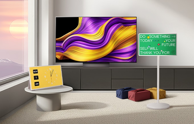 자연광이 드는 미니멀한 거실에 추상 그래픽 화면의 올레드 TV가 정면에 보이고 노란 디지털 시계 화면에 스탠바이미가 놓인 테이블, 초록색 문구 화면 스탠바이미와 선물이 배치된 장면.