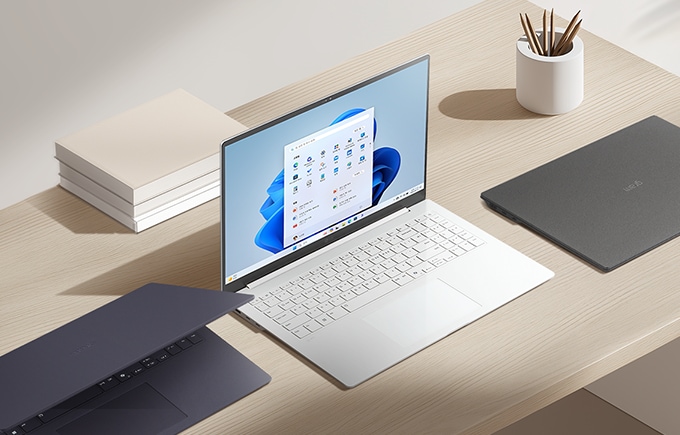 밝은 책상 위에 다양한 색상의 노트북 3대와 책, 연필꽂이가 정돈된 상태로 놓여 있습니다.