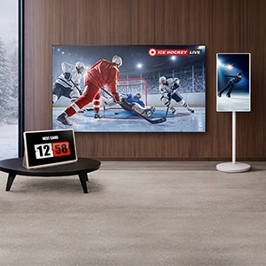 원목 벽면의 거실 공간에 아이스하키 경기 화면이 재생 중인 LG전자 벽걸이 TV가 설치되어 있고, 왼쪽 테이블 위에 시계 화면을 표시한 LG전자 스탠바이미 Go, 오른쪽에 세로 모드로 피겨스케이팅 영상이 재생 중인 LG전자 스탠바이미가 배치된 모습.