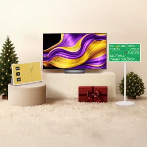 밝은 톤의 공간에 중앙 받침대 위에 보라색과 노란색 물결 무늬가 표시된 LG전자 TV가 놓여 있고, 좌측에는 노란색 시계와 조명이 켜진 크리스마스 트리가, 우측에는 초록색 문구 패널과 작은 크리스마스 트리가 배치되어 있으며, 하단에는 빨간색 리본이 달린 선물 상자가 있다.