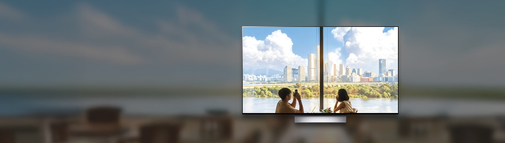 대형 TV 화면 속 창가에 앉아 강과 도심 뷰를 배경으로 사진을 찍는 남성과 맞은편에 앉아있는 여성의 모습이 보입니다.