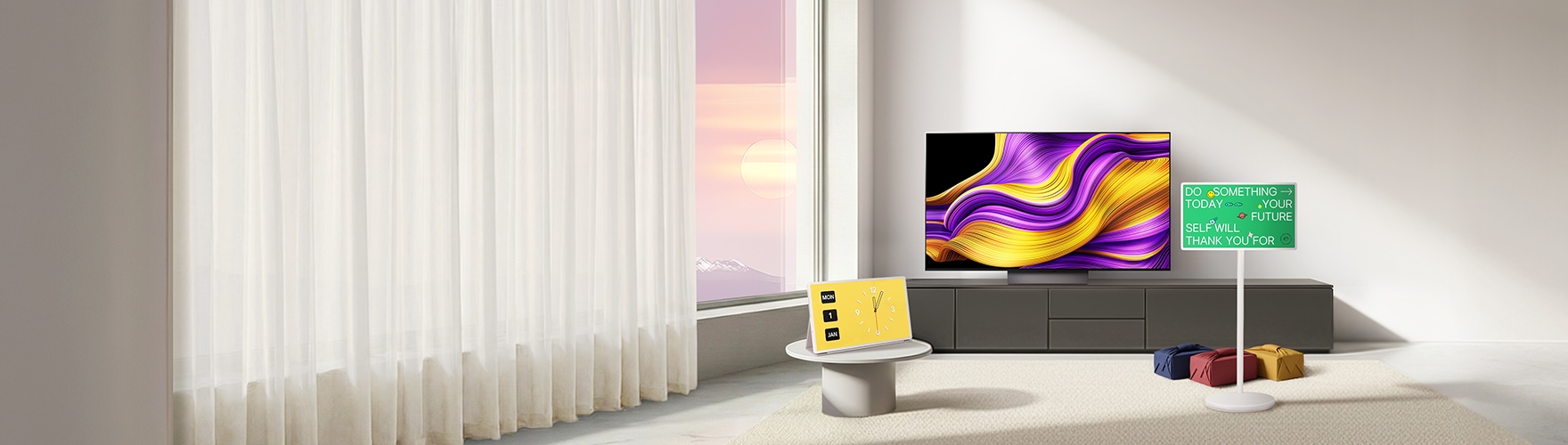 자연광이 드는 미니멀한 거실에 추상 그래픽 화면의 올레드 TV가 정면에 보이고 노란 디지털 시계 화면에 스탠바이미가 놓인 테이블, 초록색 문구 화면 스탠바이미와 선물이 배치된 장면.